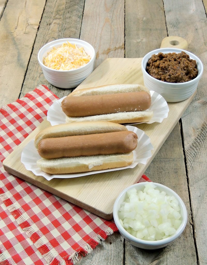 Two plain hot dogs on buns on a cutting board with bowls of chopped onions and shredded cheese on a wooden background with a red and white checkered napkin.