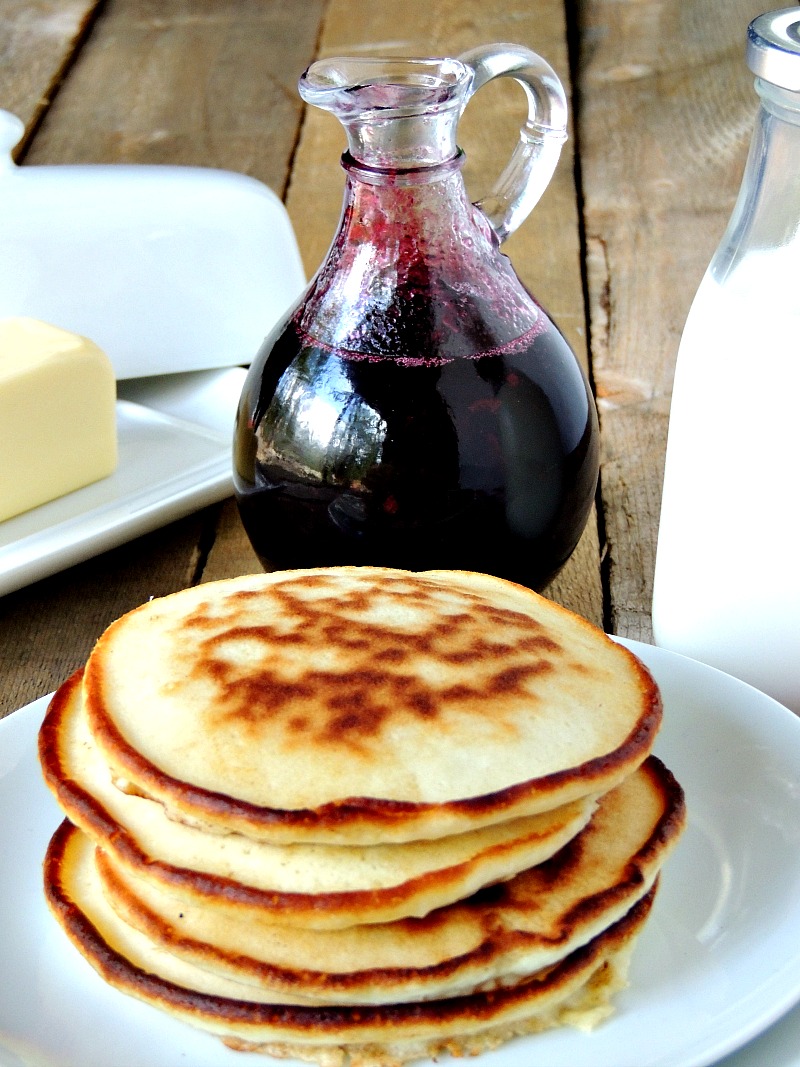 Glass bottle with Homemade Blueberry Syrup in it on a wooden table. Butter dish, glass of milk, and pancakes on a white plate with it.