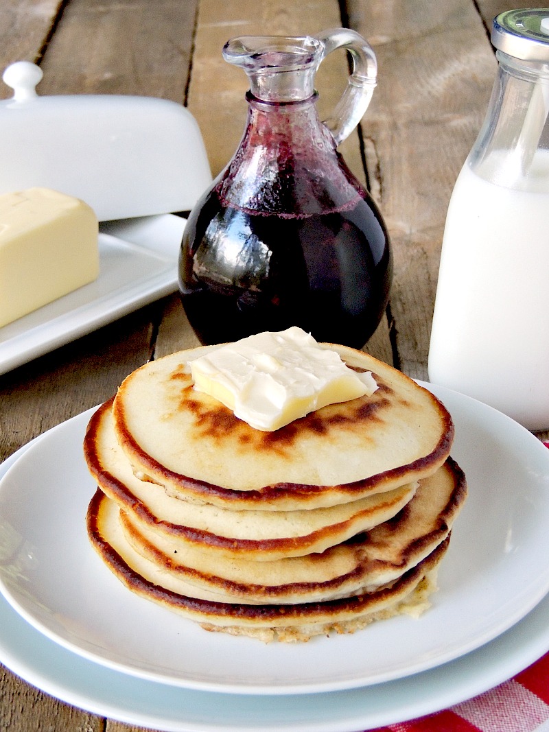 Glass bottle with Homemade Blueberry Syrup in it on a wooden table. Butter dish, glass of milk, and pancakes with butter on a white plate with it.