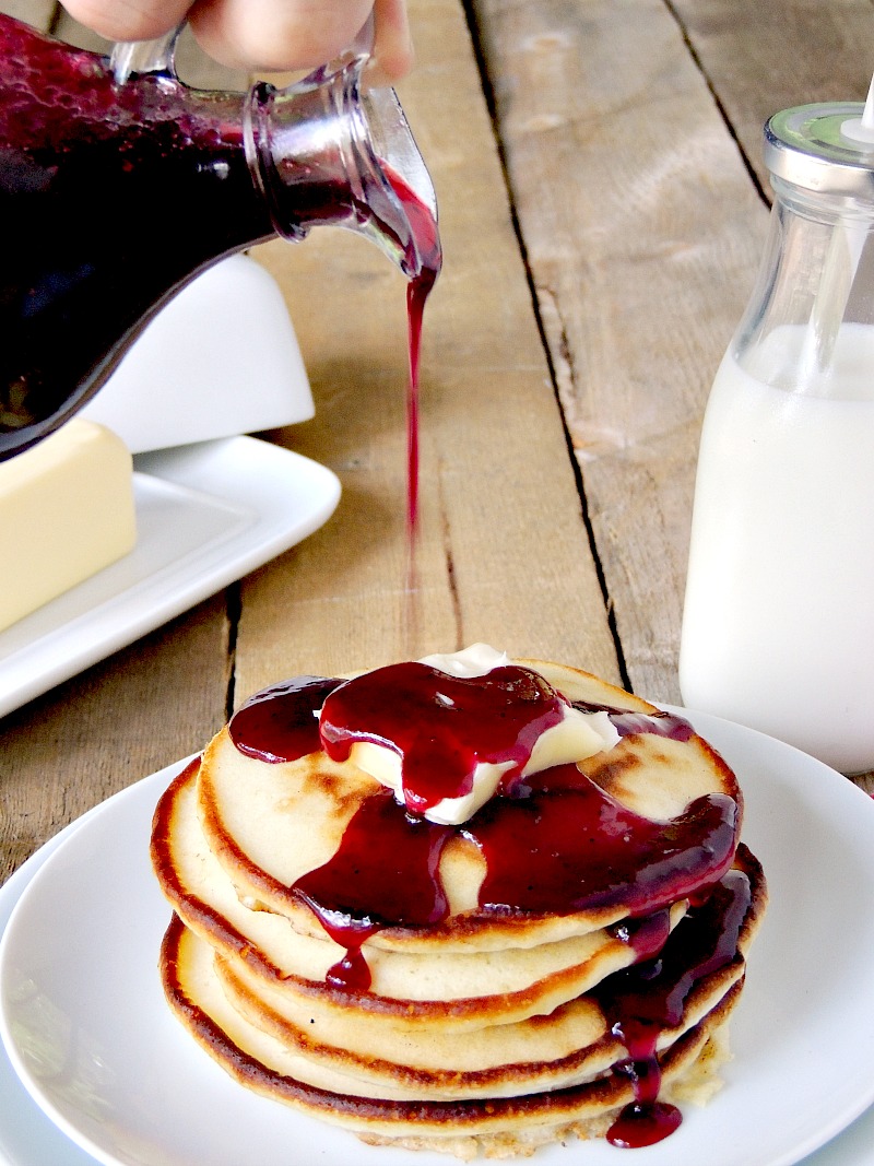 Glass bottle pouring Homemade Blueberry Syrup over pancakes with butter on a white plate on a wooden table. Butter dish and glass of milk in the background.
