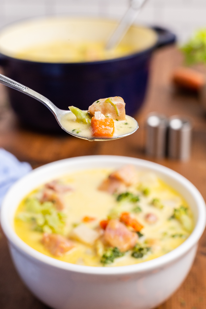 Closeup sideview of a white bowl full of Keto Cheesy Ham Chowder with a spoonful of it closer to the camera and a blue pot full blurred in the background.