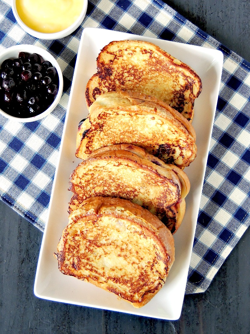 Lemon Blueberry Stuffed French Toast on a white plate with a blue and white serving platter with a small bowl of blueberries beside it on a checkered place mat.