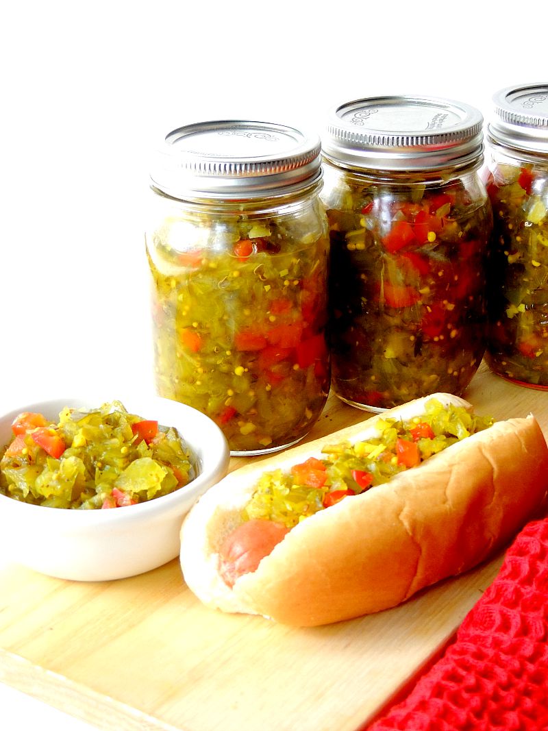 Three mason jars full of spicy pickle relish with a small white bowl full of relish and a hot dog topped with spicy pickle relish on a wooden cutting board.