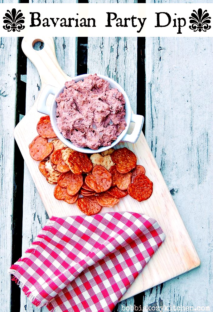 Braunschweiger Dip in a white bowl on a cutting board with pretzel chips on a wooden background with a red and white checkered napkin.