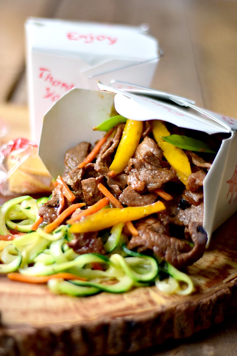 Beef Zoodle Stir Fry spilling out of a take-out container on a wooden board with chopsticks and fortune cookies