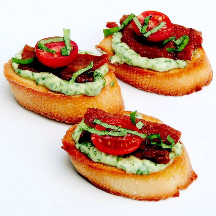 Crostini topped with basil mayonnaise, bacon, half of a grape tomato, and chopped basil, on a white background