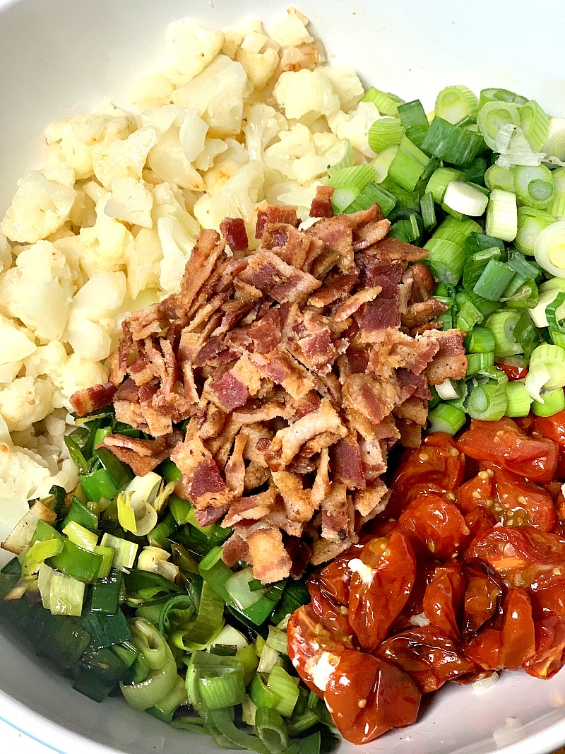 An overhead view of all the fresh and colorful ingredients needed to make the BLT cauliflower salad recipe, including crispy bacon, roasted grape tomatoes, cauliflower, sauteed leeks, and fresh chopped green onions, arranged in a large bowl before being combined, a perfect low-carb and keto-friendly side dish for any occasion.