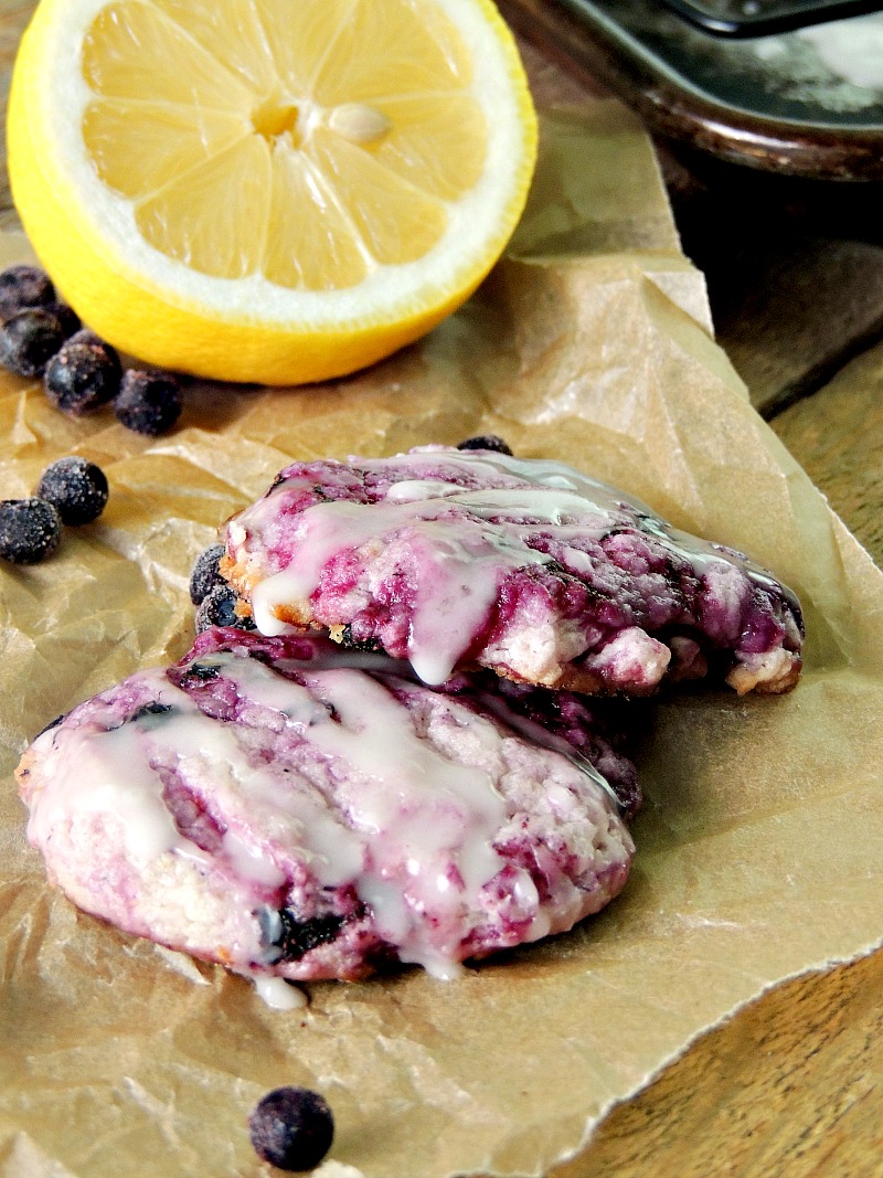 Blueberry Cheesecake Cookies with Lemon Glaze on parchment paper with blueberries and lemon in the background