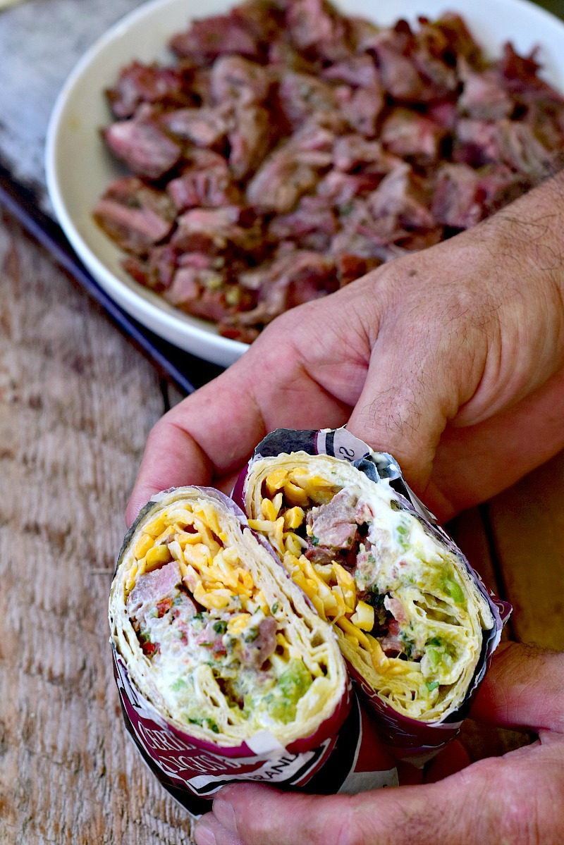 A white male presenting person holding a carne asada burrito that has been cut in half with a bowl of cooked and chopped carne asada in the background.