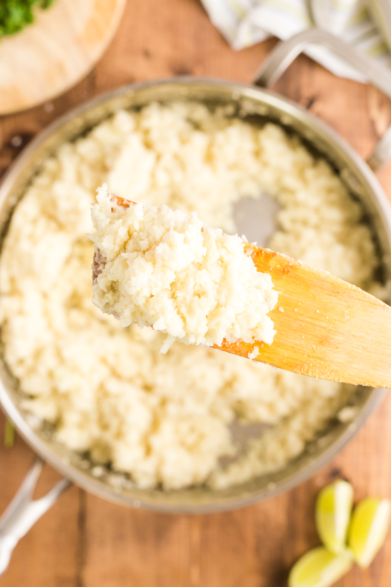 Overhead photo of Coconut Lime Cauliflower Rice in a skillet with a scoop of it on a wooden spatula closer to the camera.