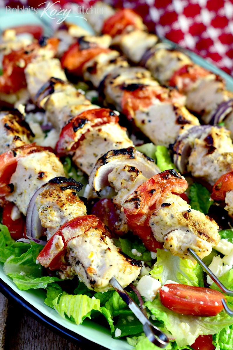 Close up of grilled chicken, tomato, and red onion kabobs on top of a Greek salad on a green serving platter.