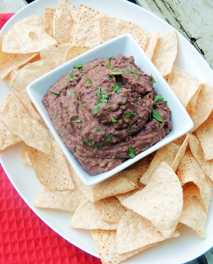 Bean dip in a square while bowl on a white platter with tortilla chips. Red towel and wooden background.