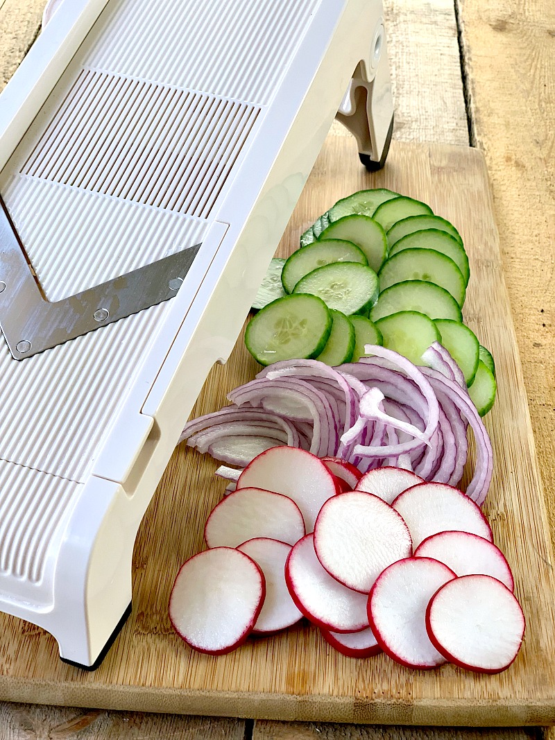Sliced cucumber, red onion, and radish on a wooden cutting board next to a madolin.