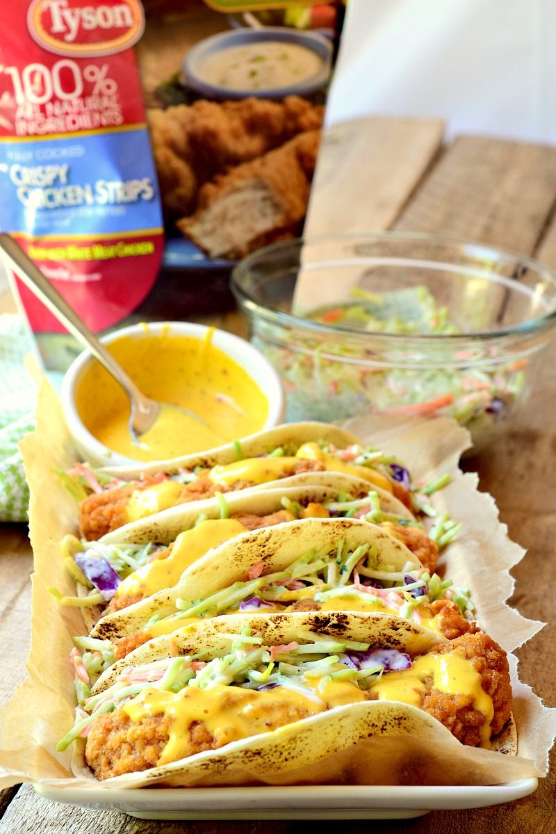 This photo shows four tacos made with honey mustard chicken and broccoli slaw, arranged on a parchment-lined baking sheet. A bag of frozen chicken tenders, a small bowl of the honey mustard sauce, and a bowl of remaining broccoli slaw are also shown on a wooden table next to the baking sheet.