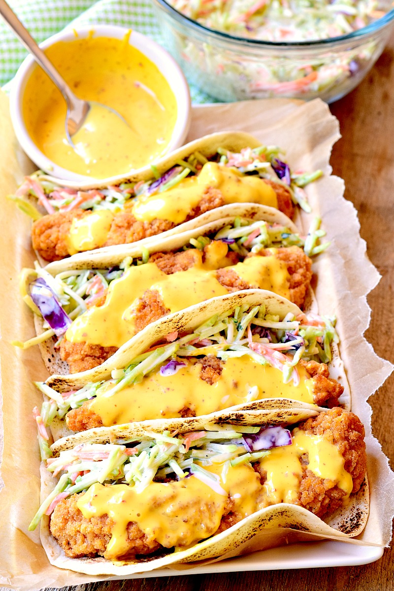 This photo shows four Honey Mustard Chicken Tacos arranged on a parchment-lined baking sheet. A small bowl of the honey mustard sauce and a bowl of remaining broccoli slaw are also shown on a wooden table next to the baking sheet.