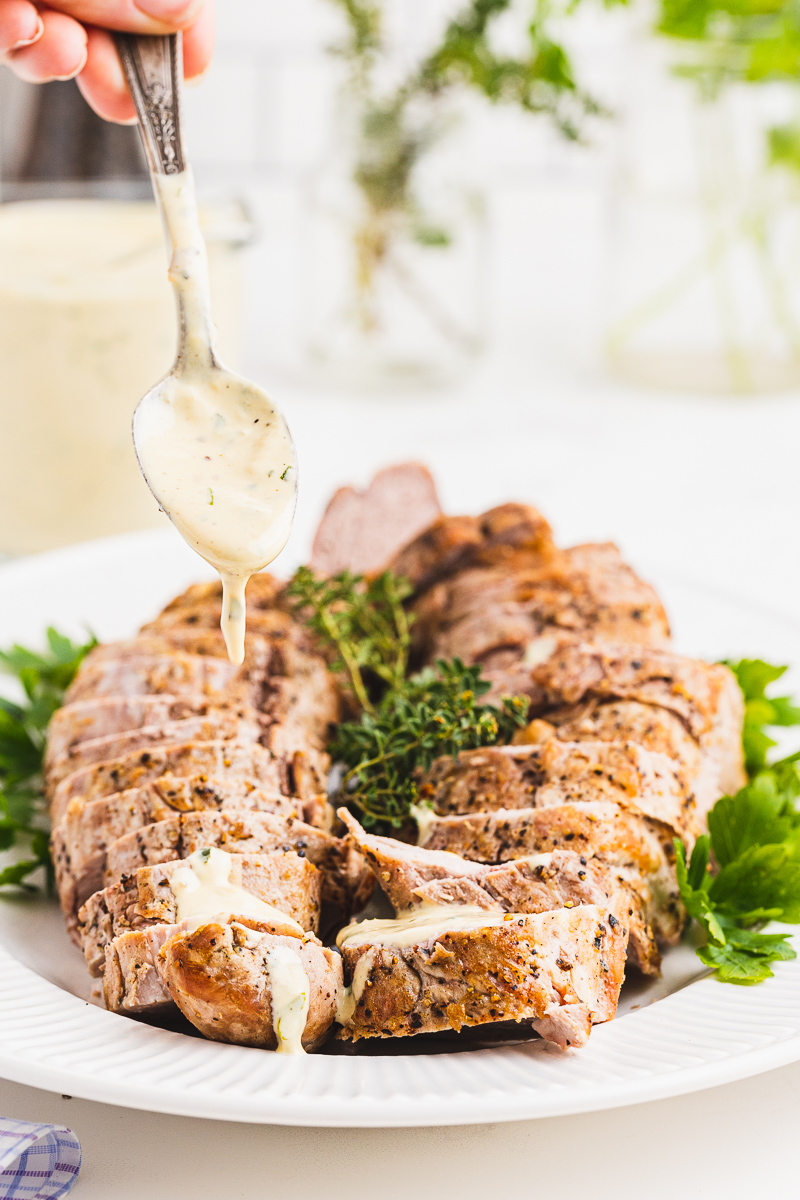 Closeup of a spoon drizzling the herb mustard sauce over 2 cooked pork tenderloins.