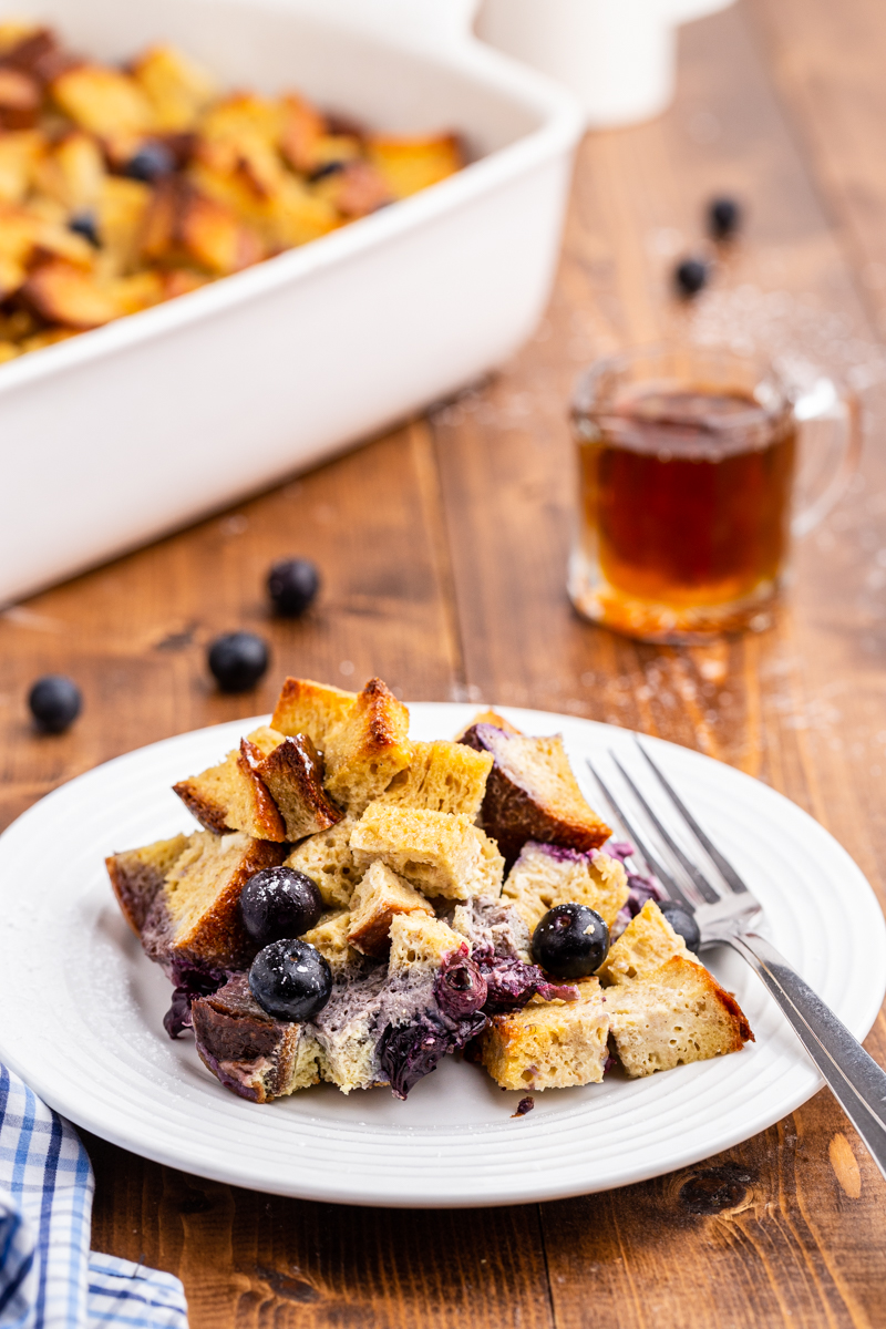 A serving of Keto Blueberry French Toast Bake on a white plate with the baking dish containing the remaining casserole in the background.