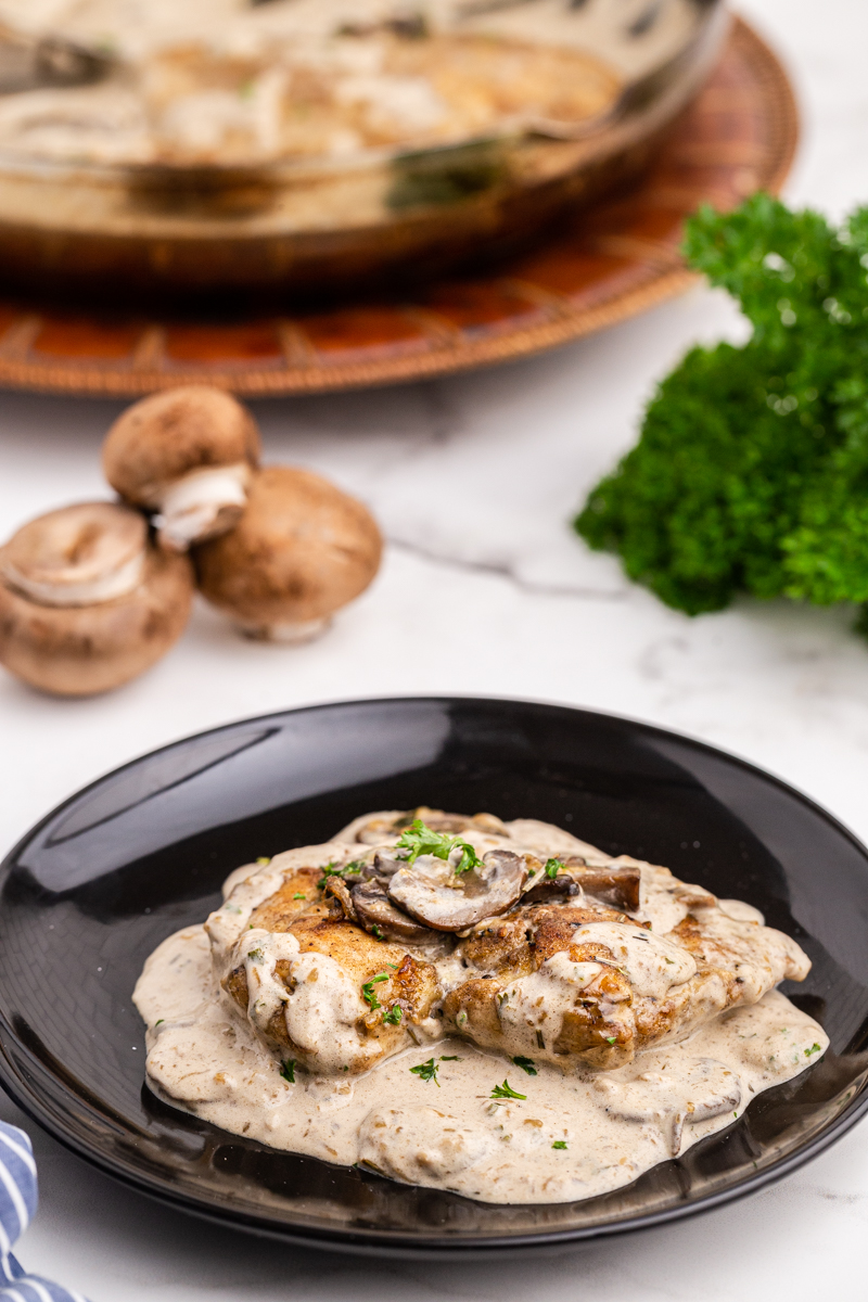 Closeup of a serving of Chicken with Mushroom Garlic Sauce on a black plate with fresh mushrooms in the background.