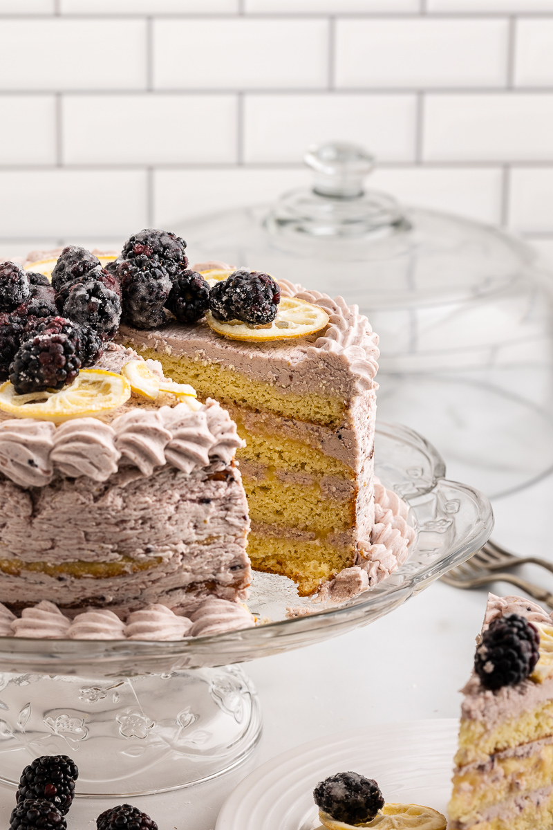 Photo of a Keto Lemon Cake with Blackberry Mascarpone Frosting on a glass cake stand with a slice cut out.