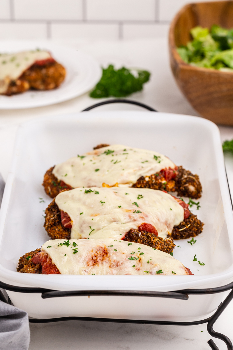 Keto Sheet Pan Chicken Parmesan in a white baking dish with a serving on a white plate in the background.