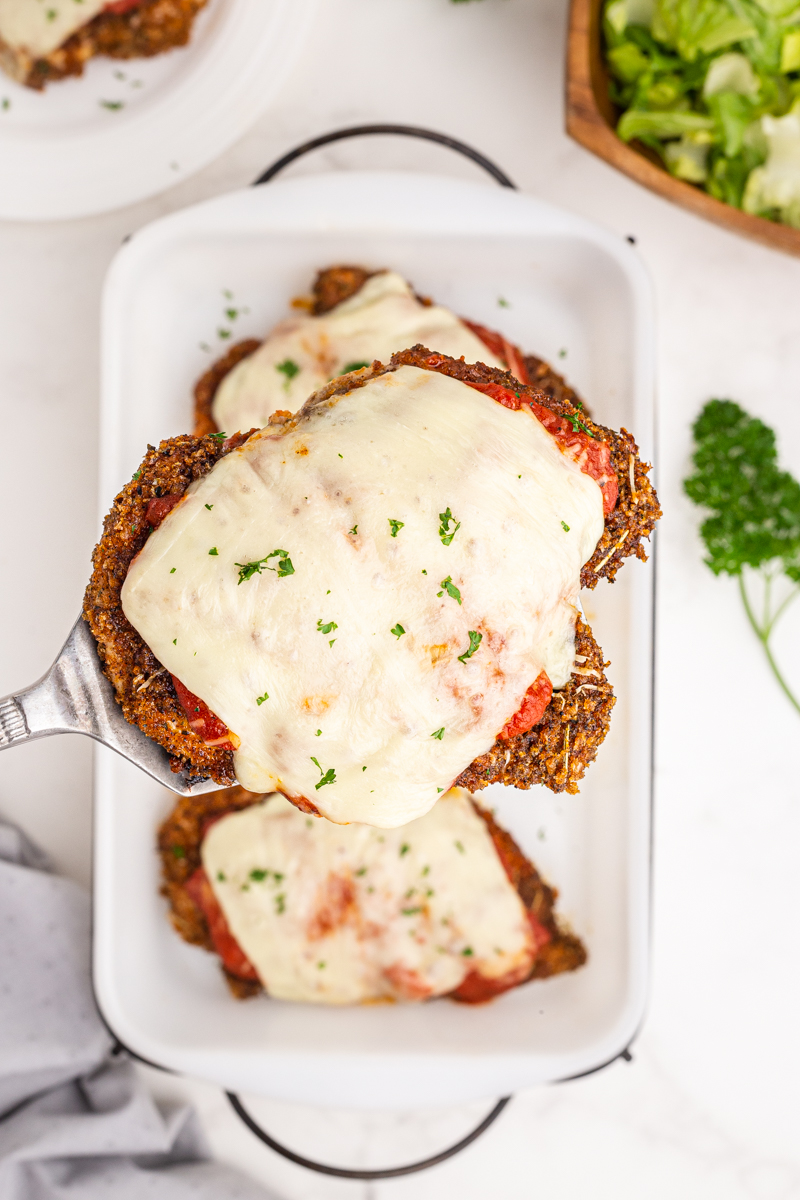 Overhead photo of a single serving of Keto Sheet Pan Chicken Parmesan on spatula above a baking dish.