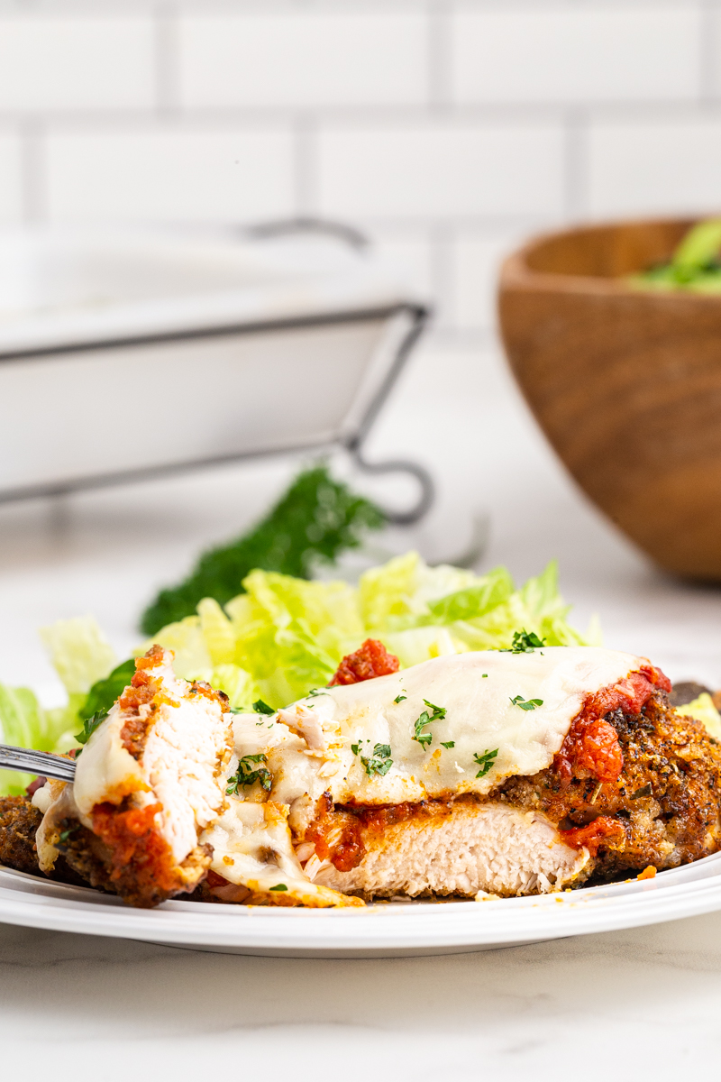 Closeup of a single serving of Keto Sheet Pan Chicken Parmesan on a white plate with a bite on a fork to show the inside of the chicken.