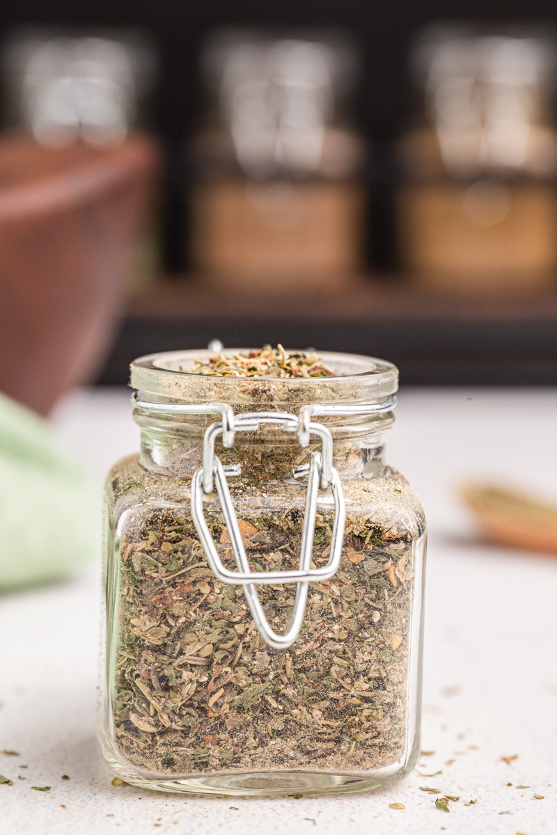 Closeup photo of Homemade Italian Seasoning in a glass spice jar  on a white table with a wooden spice rack in the background.