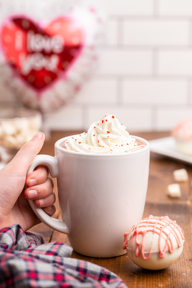 Photo of hot chocolate in a white mug with a Keto Valentines Hot Chocolate Bomb next to it on a wooden table.