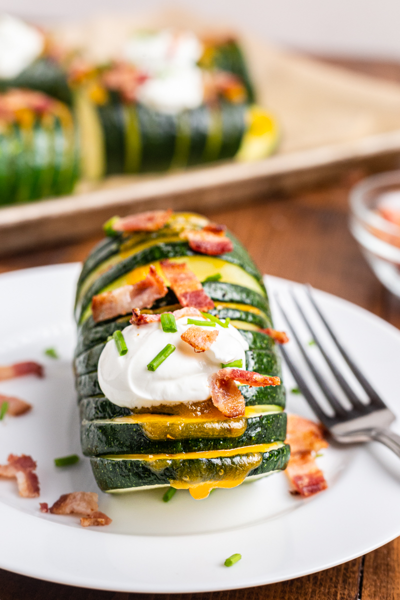 One serving of Hasselback zucchini on a white plate topped with sour cream and bacon crumbles.