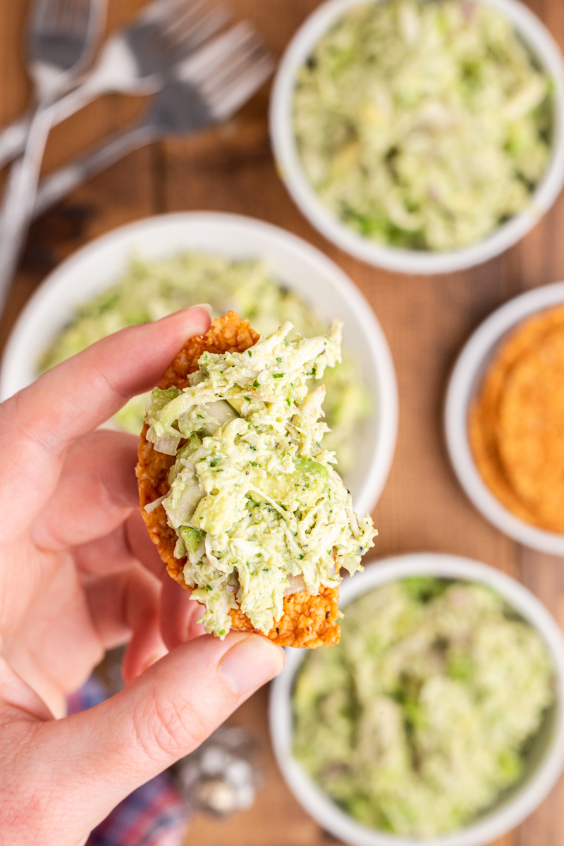 Photo of a hand holding a keto cracker with Avocado Ranch Chicken Salad on it over a wooden table with several bowls full of the chicken salad in the background.