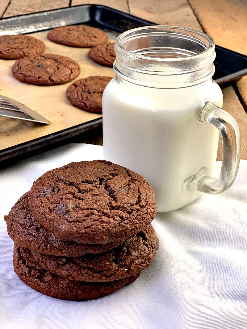 Photo of Keto Double Chocolate Chip Cookies on a white plate with a mason jar of milk next to the plate and cookies on a cookie sheet in the background.