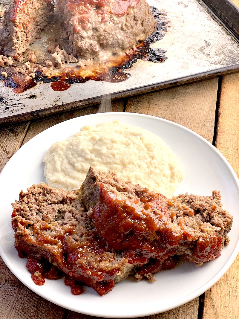 Photo of Keto Homestyle Meatloaf on a white plate with mashed cauliflower on a wooden table with the rest of the meatloaf on a sheet pan in the background.