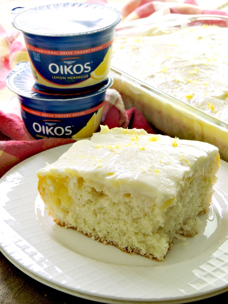Sunshine Lemon Poke Cake in a glass dish on a wooden table with a yellow and red plaid table runner and Oikos Lemon Meringue yogurt.