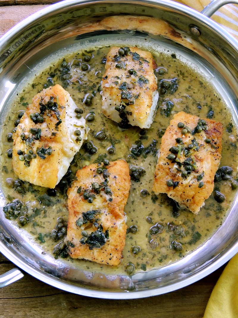 Overhead view of a silver skillet filled with four perfectly cooked cod filets, each one adorned with a generous drizzle of tangy lemon white wine sauce. The filets are beautifully garnished with scattered capers, adding a touch of saltiness and visual appeal.