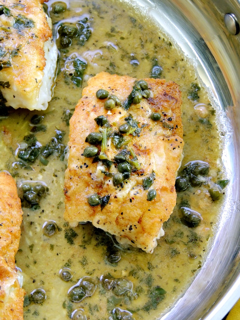 Close-up view of a perfectly cooked cod filet in a skillet, showcasing its golden-brown crust and delicate flaky texture. The filet is adorned with a drizzle of tangy lemon white wine sauce, and capers are studded across the top, adding a touch of saltiness and visual appeal.