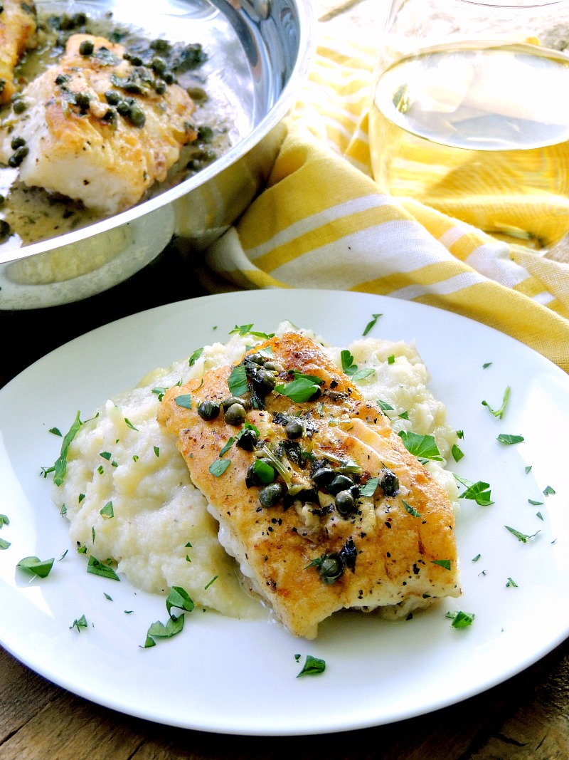 A mouthwatering dish of pan roasted cod filet served on a bed of creamy mashed cauliflower, topped with a tangy lemon white wine sauce and scattered capers. The plate sits elegantly on a rustic wooden table, accompanied by a pan filled with more succulent filets. A charming yellow and white striped towel adds a delightful touch to the background.