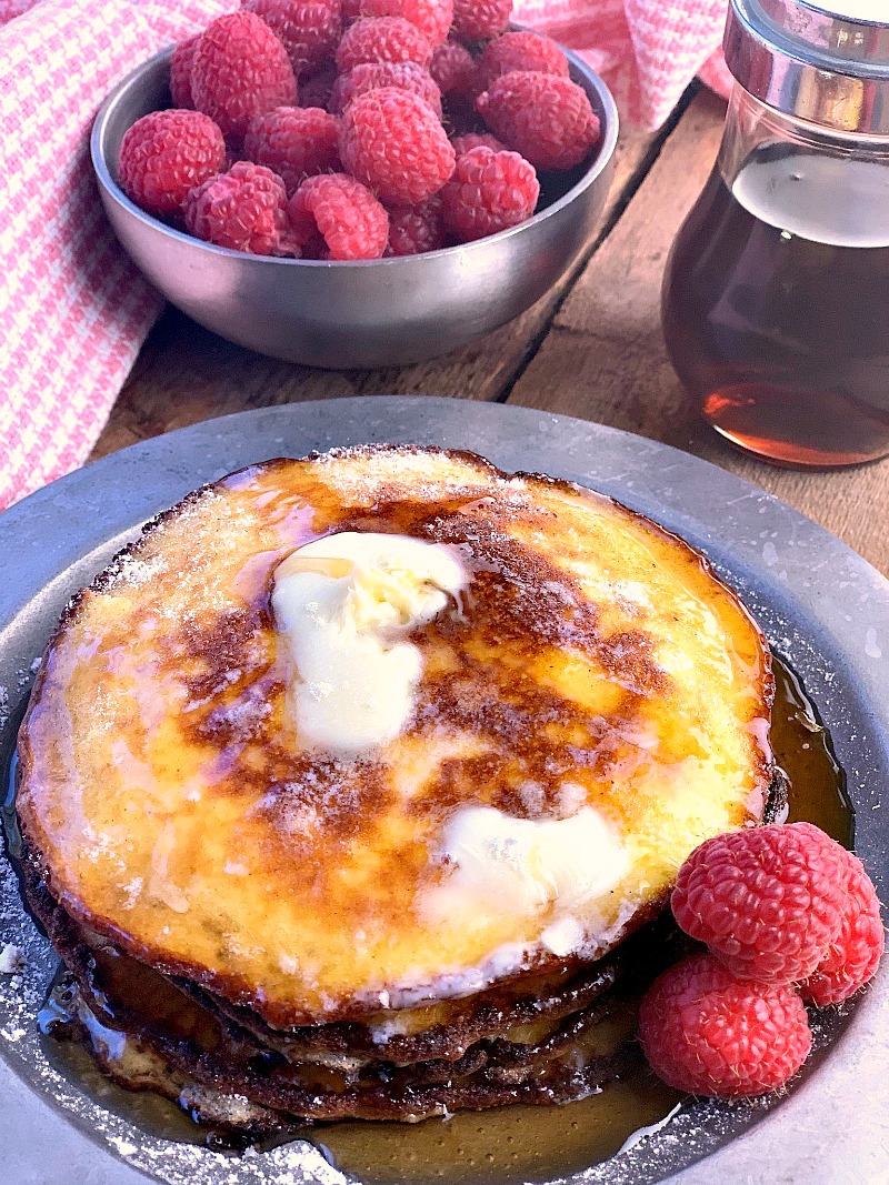 Photo of Cream Cheese Pancakes on a metal plate with butter and syrup on them and berries in a bowl on the side.