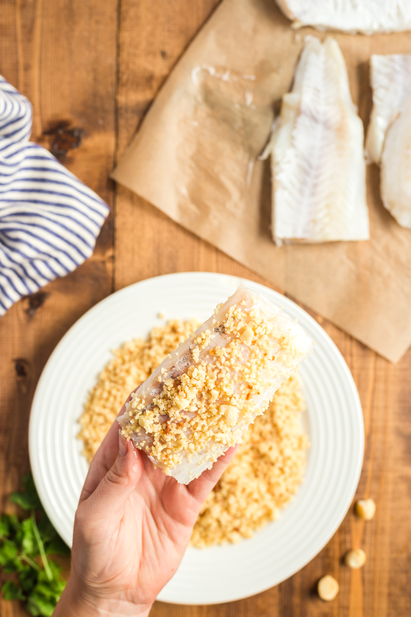 A white female presenting hand holding a macadamia but crusted cod filet.