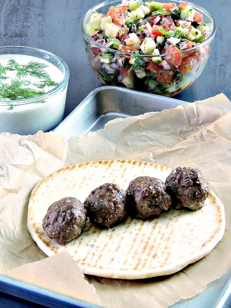 Gyro meatballs on a pita round on a parchment paper lined baking sheet with tzatziki and cucumber relish in bowls.