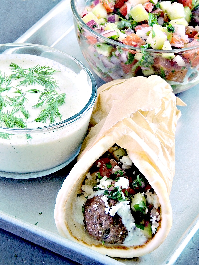 Gyro meatballs sandwich wrapped in parchment paper on a baking sheet with tzatziki and cucumber relish in bowls.