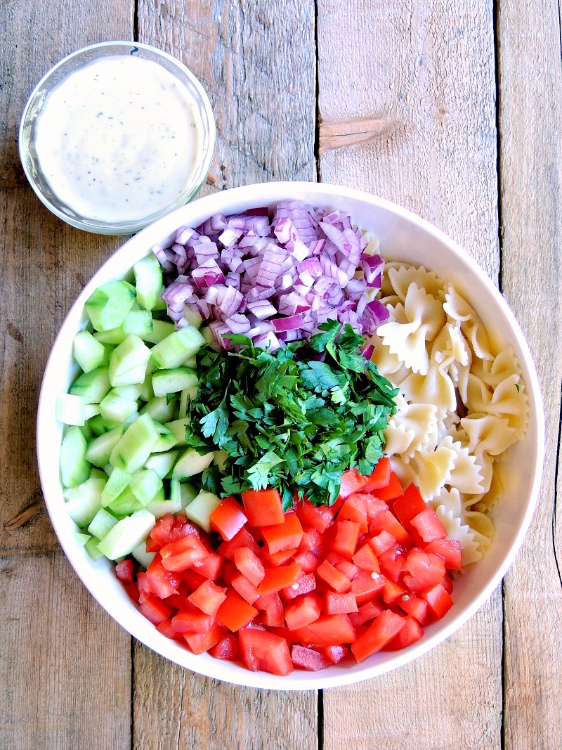 Ingredients for the Mediterranean Pasta Salad in a bowl with a with a small bowl of salad dressing next to it on a wooden background