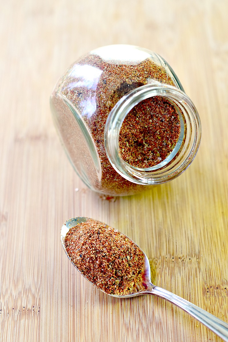 An image showcasing a glass spice jar filled with Homemade Mexican Spice Blend, placed on its side on a wooden table. A spoon lies beside the jar, filled with the flavorful spice mixture.