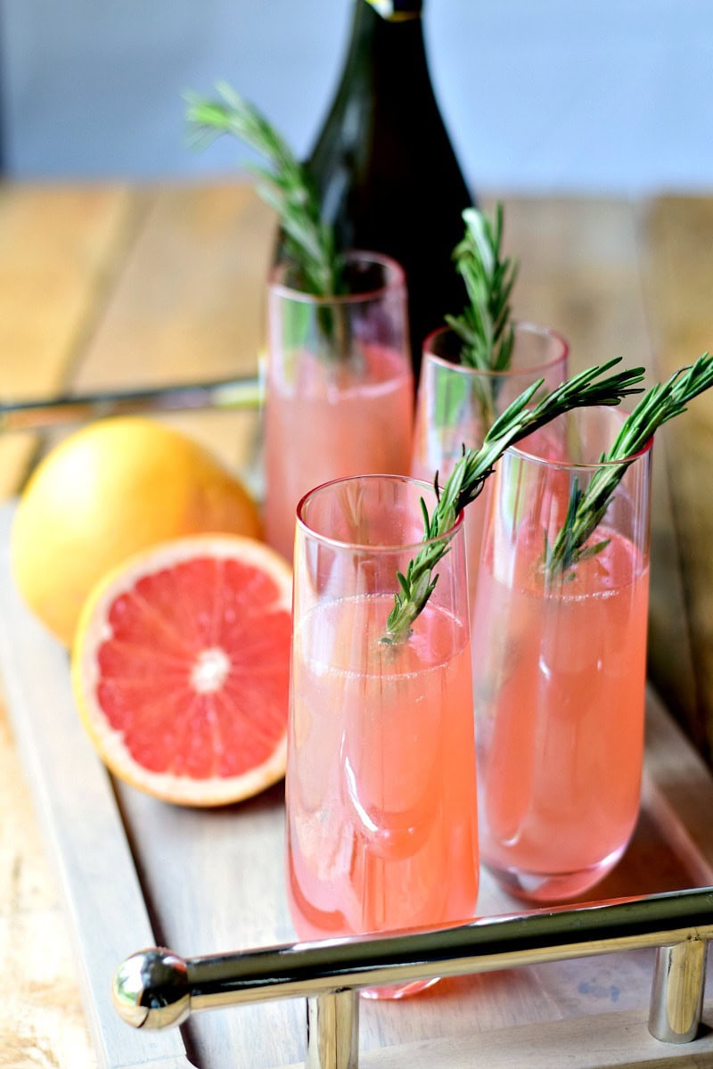 Several sugar-free roasemary pink grapefruit mimosas on a wooden tray with a grapefruit cut in half and a bottle of chgampagne next to them.