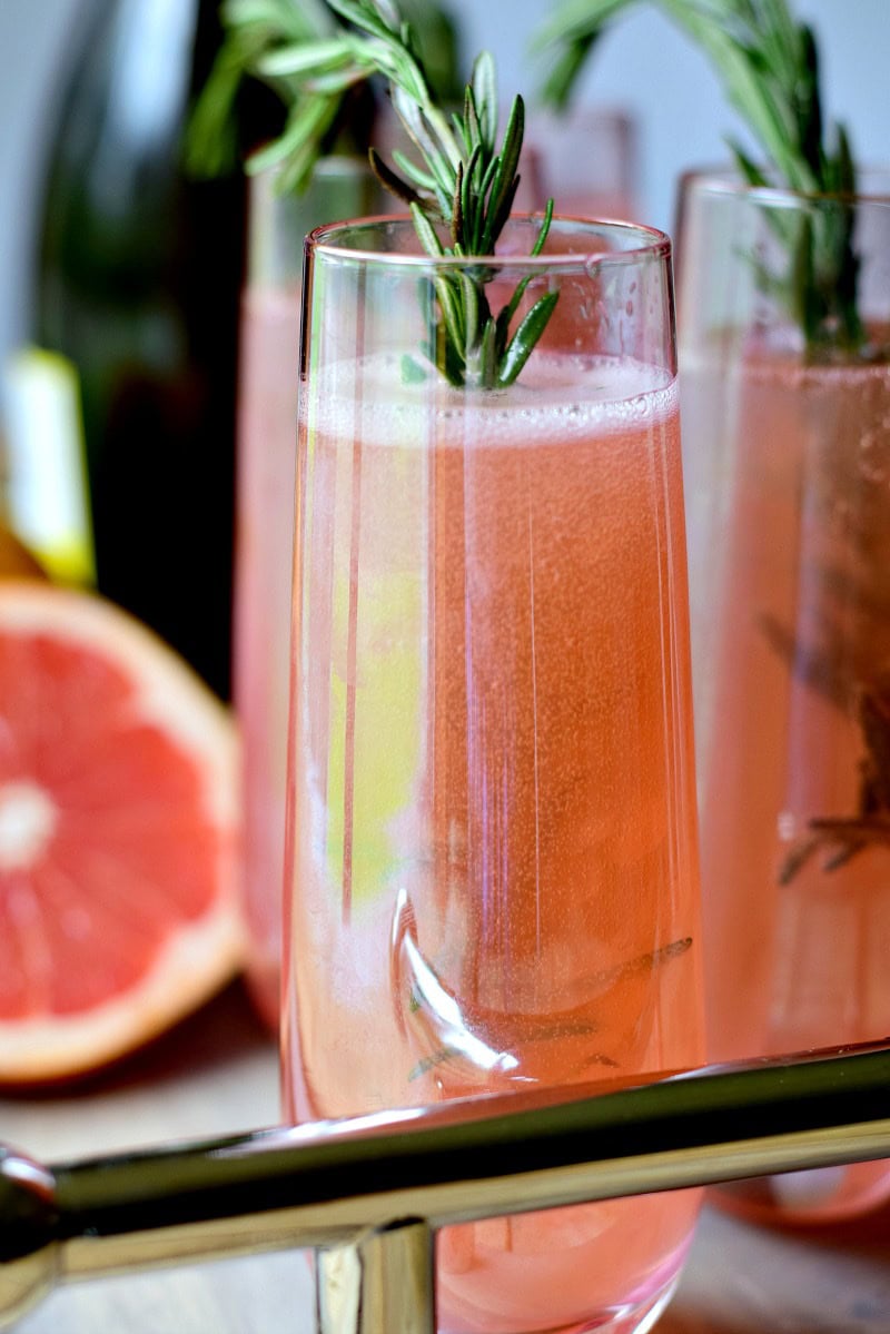 Several glasses of sugar-free roasemary pink grapefruit mimosas on a wooden tray with sprigs of rosemary in the glasses.