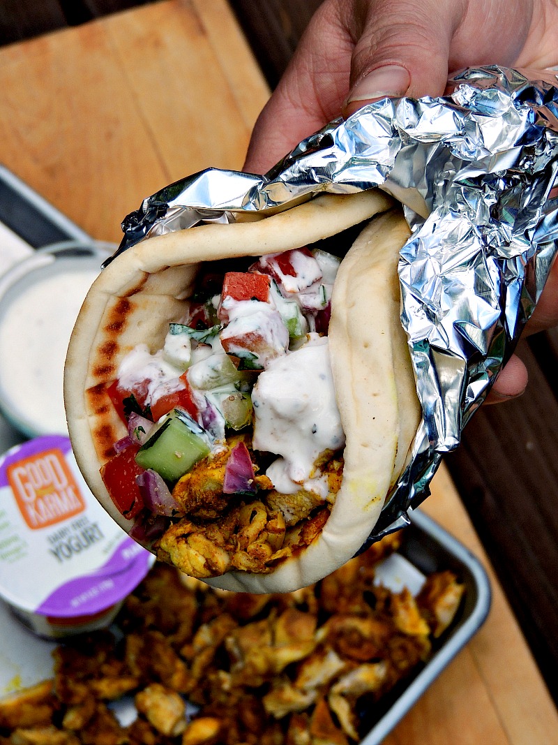 Delicious Sheet Pan Chicken Shawarma wrapped in a pita and topped with raita, held by a hand. The pita is filled with flavorful marinated chicken, accompanied by a refreshing yogurt sauce. The meal is wrapped in foil. In the background, a sheet pan is visible with the remaining ingredients from the recipe.