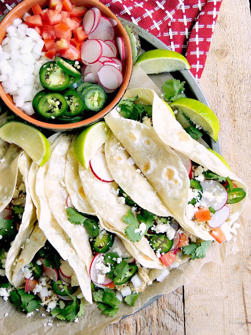 Overhead view of a serving plater full of these green chile chicken tacos with a bowlful of garnish like chopped tomato and onion, and sliced radish and jalapeno.