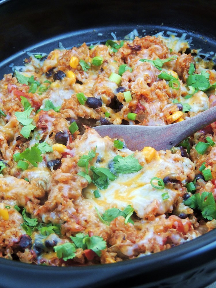 Chicken Enchilada Quinoa in a slow cooker, with a wooden spoon scooping it out. You can see the corn, tomatoes, and black beans and the quinoa is topped with melted cheese, fresh chopped green onions, and cilantro.