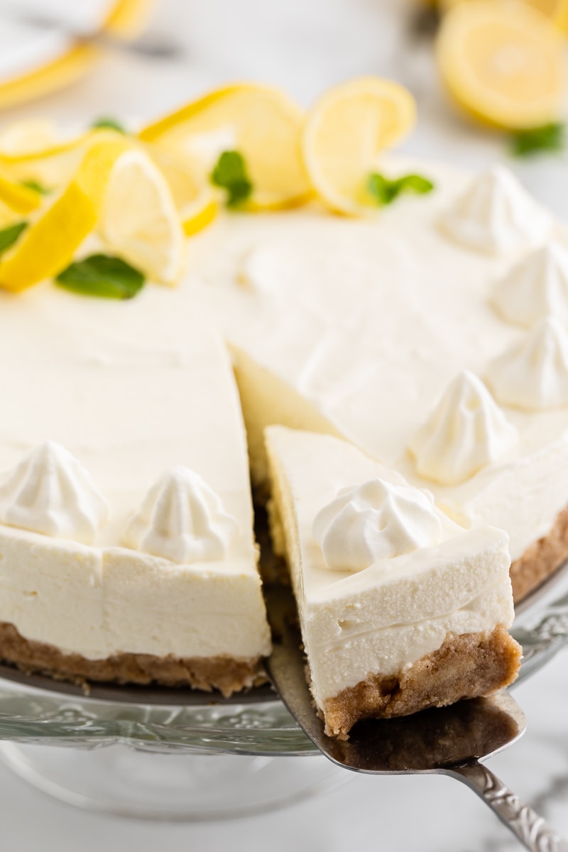 Keto no-bake lemon cheesecake on a glass cake stand with one slice cut out on a silver pie cutter.