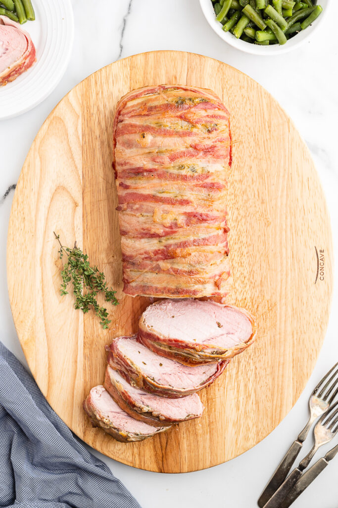 Overhead photo of a bacon wrapped honey mustard glazed pork loin sliced on a wooden cutting board.