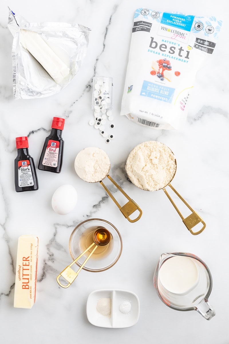 An overhead view of all of the ingredients needed to make keto cheesecake mummy cookies on a white marble counter.